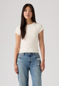 Νεαρή γυναίκα με μακριά σκούρα μαλλιά, ντυμένη με λευκό κοντομάνικο πουκάμισο με μικρό μοτίβο και ψηλόμεσο μπλε τζιν με ελαφρύ ξεφλούδισμα.