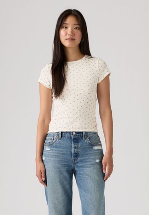 Νεαρή γυναίκα με μακριά σκούρα μαλλιά, ντυμένη με λευκό κοντομάνικο πουκάμισο με μικρό μοτίβο και ψηλόμεσο μπλε τζιν με ελαφρύ ξεφλούδισμα.