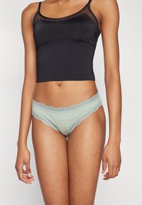 Hellgrüne Unterwäsche mit Spitze und weichem Stoff, kombiniert mit einem schwarzen, figurbetonten Tanktop mit transparenten Akzenten und verstellbaren Trägern.