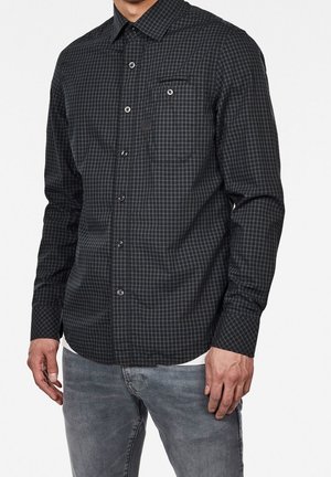 Homme portant une chemise à carreaux foncée à boutons avec poche poitrine et un jean gris clair, debout devant un fond blanc uni.