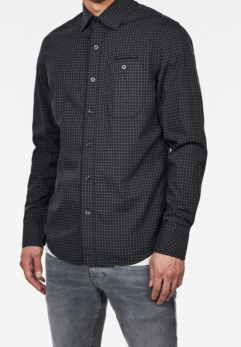 Homme portant une chemise à carreaux foncée à boutons avec poche poitrine et un jean gris clair, debout devant un fond blanc uni.