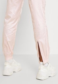 Ljusrosa träningsbyxor i glänsande tyg, med siddragkedjor och elastiska muddar, kombinerat med vita chunky sneakers.