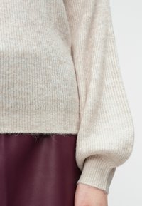 Beżowy sweter z prążkowanej dzianiny z rękawem zakończonym mankietem spoczywającym nad bordową skórzaną spódnicą, ukazując tekstury i kontrast materiałów na ramieniu i talii.