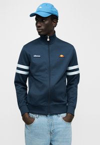 Σκούρο μπλε ζακέτα με φερμουάρ, λευκές ρίγες στα μανίκια, κεντημένο λογότυπο και ψηλό γιακά, σε συνδυασμό με ανοιχτό γαλάζιο καπέλο.