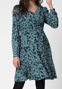 Teal-kangasta valmistettu kukkamekko mustilla kukilla. Ominaisuuksiin kuuluu rypistetty rintaosa, pitkät hihat ja levenevähelma. Kauluksessa on nappeja.