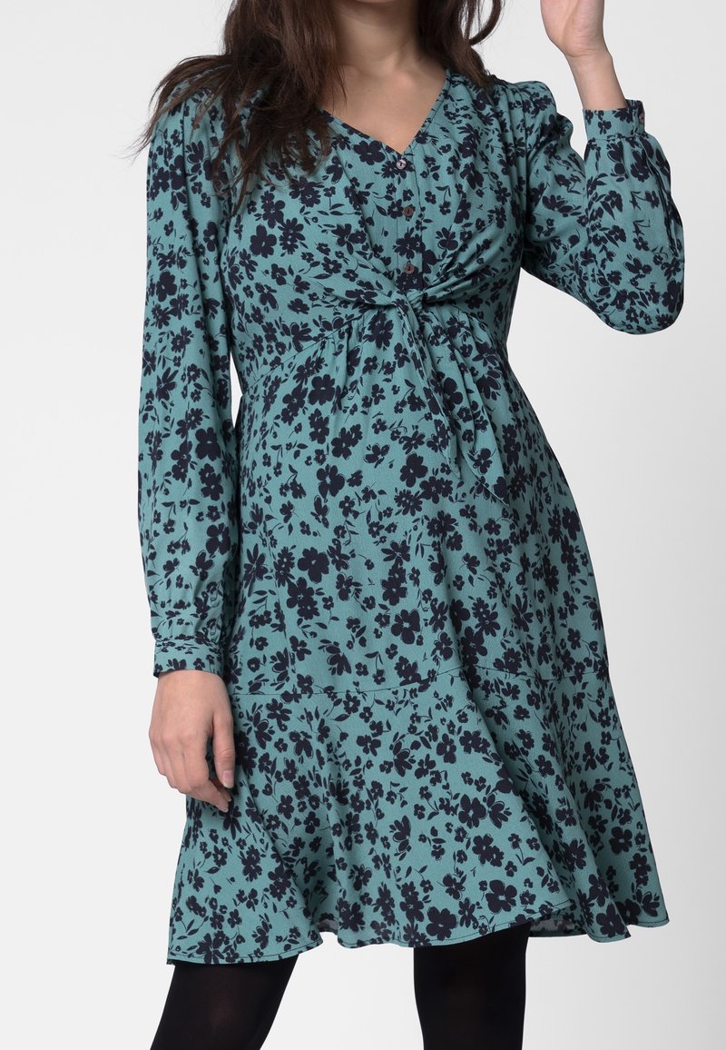 Teal-kangasta valmistettu kukkamekko mustilla kukilla. Ominaisuuksiin kuuluu rypistetty rintaosa, pitkät hihat ja levenevähelma. Kauluksessa on nappeja.