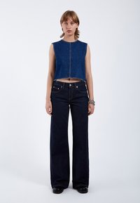 Chaleco de mezclilla recortado con dobladillo deshilachado, de color azul claro y sin mangas, combinado con jeans de pierna ancha de color azul oscuro que cuentan con bolsillos frontales y botones.