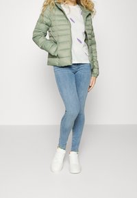 Lys grøn pufferjakke med lynlås, kombineret med en hvid t-shirt med lilla bladbånd og blå skinny jeans. Hvide sneakers.