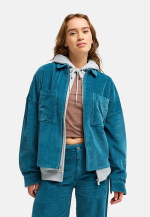Junge Frau trägt eine türkisfarbene Cordjacke und Hose über einem grauen Hoodie und braunem Crop-Top, stehend vor weißem Hintergrund.