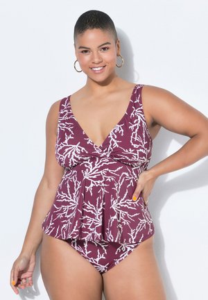 Donna sorridente con capelli corti che indossa un costume tankini bordeaux e bianco con motivo, in piedi con la mano sul fianco su sfondo bianco.