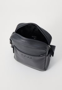 Pelēka ādas crossbody soma ar r�āvējslēdzēja aizdari, priekšējo kabatu un regulējamu siksnu. Iezīmēta ar iepresētu logo priekšpusē.