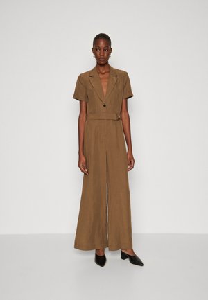Tuta in lino marrone con vita aderente, maniche corte, scollo a V profondo e pantaloni a gamba larga. Completa il look un paio di tacchi neri con punta a punta.