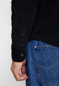 Czarna koszula z kordurowego materiału z guzikowym mankietem, w połączeniu z niebieskimi dżinsami z subtelnym detalem przeszycia na tylnej kieszeni.