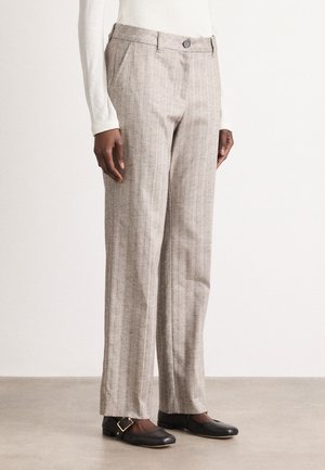 Persoon in beige gestreepte recht-toe-rechtdoor broek, gedragen met zwarte platte schoenen met gouden gespdetails.