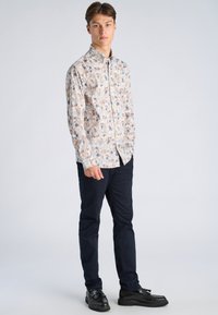 Noor mees seisab, kandes beežikat lillemustriga nööbitavat särki, tumelisi siniseid pükse ja musti narmastega loafereid, taustaks hele ühevärviline sein.