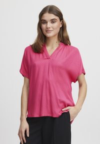 Blusa rosa a maniche corte con scollatura a V, caratterizzata da un motivo testurizzato e una vestibilità comoda, abbinata a pantaloni neri.