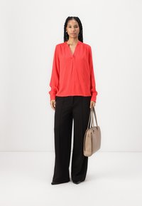 Blusa rossa a maniche lunghe con scollatura a V e una sottile texture verticale, abbinata a pantaloni neri a gamba larga con vita alta, mentre tiene una borsa beige.