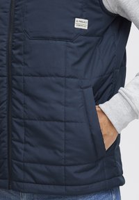 Dunkelblau gesteppte Weste mit Taschen, mit Reißverschluss und einem kleinen eingenähten Etikett. Graue Pulloverü rmel sind am Kragen sichtbar.