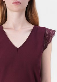 Bordový top s výstřihem ve tvaru V a krátkými rukávy. Levý rukáv zdobí propracované krajkové detaily. Materiál má hladký, mírně texturovaný povrch.