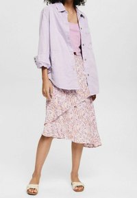 Camicia button-up viola chiaro con maniche arrotolate sopra una canotta rosa, abbinata a una gonna a lunghezza ginocchio con un motivo nei toni del rosa e del giallo.