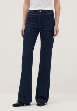 Donkerblauwe jeans met wijde pijpen en hoge taille, gedragen met zwarte laarzen met vierkante neus en een wit, ingestopt shirt, getoond van halverwege de torso tot aan de voeten.