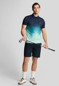 Navyblauwe polo met korte mouwen en een verloop van turkoois, gecombineerd met navy korte broek en witte sportschoenen, terwijl hij een golfclub vasthoudt.