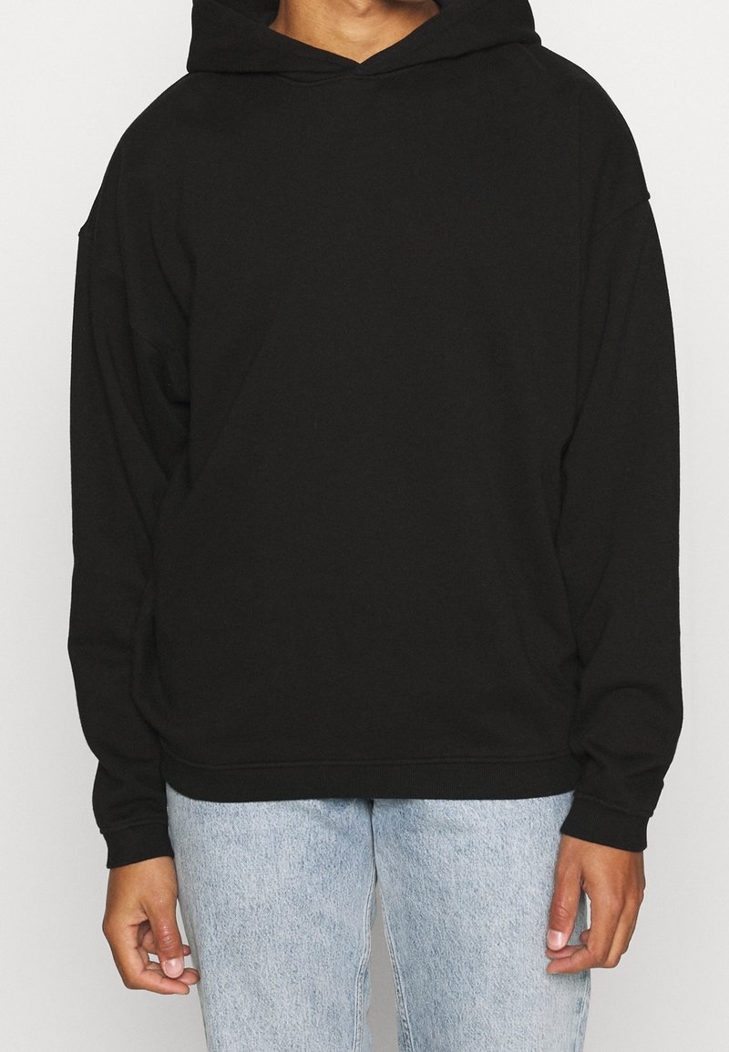 Personne portant un sweat à capuche noir uni et un jean bleu clair, debout devant un fond neutre.