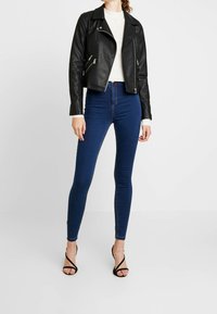 Chaqueta de biker corta de cuero negro con cremalleras plateadas, combinada con jeans skinny azul oscuro y tacones negros de tiras.