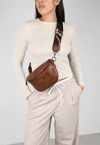 Bolso de piel marrón tipo bandolera con forma curva, que cuenta con un cierre de cremallera y una correa estampada en tonos terrenales. Se lleva a la cintura.