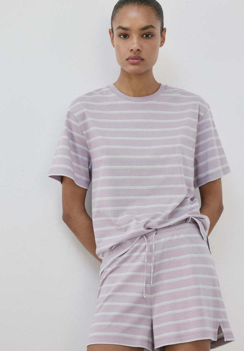 Jeune femme debout contre un mur blanc, portant un t-shirt à manches courtes à rayures horizontales violet clair et blanc et un short assorti.