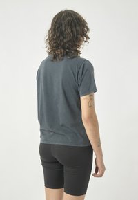 Graues Baumwoll-T-Shirt mit lässiger Passform und kurzen Ärmeln, von hinten gezeigt. Getragen mit schwarzen, eng anliegenden Shorts. Auffälliges Tattoo am Arm.