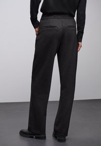 Pantalons noirs amples avec une finition texturée subtile, dotés de deux poches arrière et portés avec des chaussures noires.
