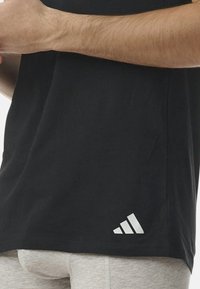 Svart bomullströja med korta ärmar och normal passform, med en vit adidas-logotyp på nedre vänstra sidan. Slät textur.