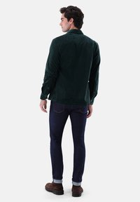 Dark green corduroy shirt with a collar, buttoned cuffs, and back yoke detail, paired with dark blue jeans and brown shoes.