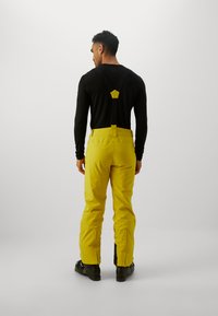 Salopete galbene impermeabile cu bretele ajustabile și fermoare laterale. Purtați peste o bluză neagră cu mânecă lungă, având un design simplu și funcțional.