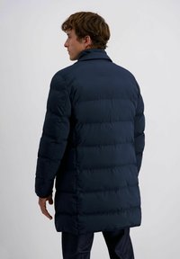 Homme portant un manteau matelassé bleu marine vu de dos, se tenant contre un fond gris clair uni.