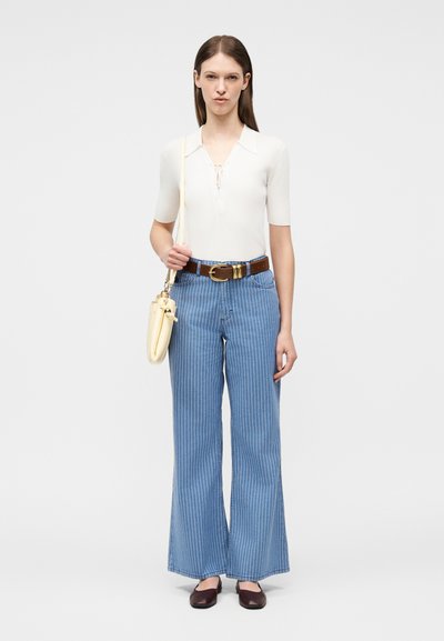 Γυναίκα που φοράει λευκό πλεκτό μπλουζάκι με κοντά μανίκια, καφέ ζώνη, μπλε φαρδιά τζιν με ρίγες, σκούρα καφέ παπούτσια και κρατάει ένα κρεμ τσάντα ώμου.