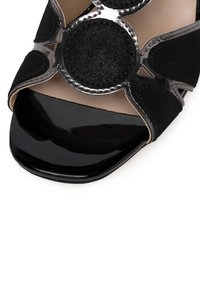 Tacón alto negro con punta abierta que presenta materiales de ante y cuero liso, acentos circulares decorativos y un acabado negro brillante en la punta.