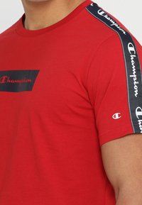 Camiseta de algodón roja con una banda de logo rectangular negra, que presenta el texto blanco "Champion". Incluye un detalle de cinta de marca en blanco y negro en las mangas.