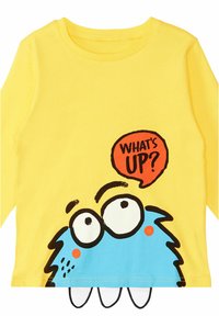 Gelbes Langarm-T-Shirt aus Baumwolle, mit einem blauen Monster-Graphic, das große Augen hat und einer roten Sprechblase mit der Aufschrift "WAS GIBT'S?".