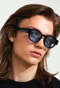 Black sunglasses with thick frames and round lenses featuring a blue tint, set against a neutral background. Hair softly frames the face.