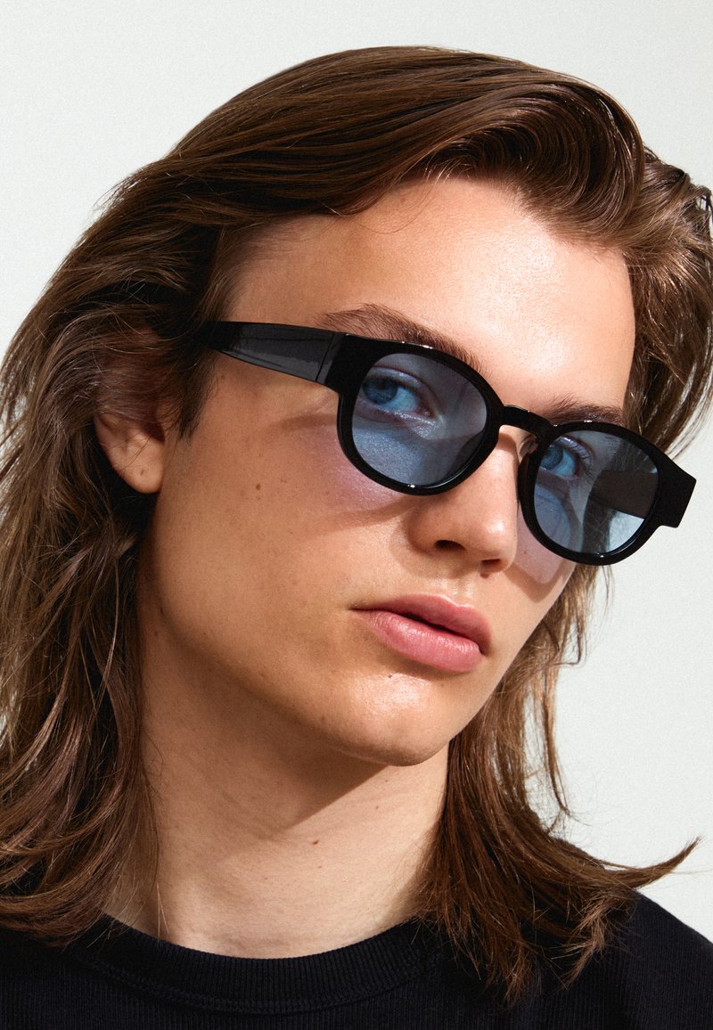 Black sunglasses with thick frames and round lenses featuring a blue tint, set against a neutral background. Hair softly frames the face.