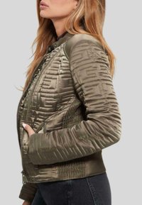 Veste bomber matelassée vert olive avec fermeture éclair, présentant des panneaux texturés et des poches latérales. Matière lisse et rembourrée avec une coupe ajustée.