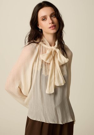 Nuori nainen, jolla on pitkät tummat hiukset, pukeutuneena läpikuultavaan kermanväriseen puseroon, jossa on suuri rusettikaulus, ja ruskeisiin housuihin, yksinkertaisen vaaleanbeigen taustan edessä.