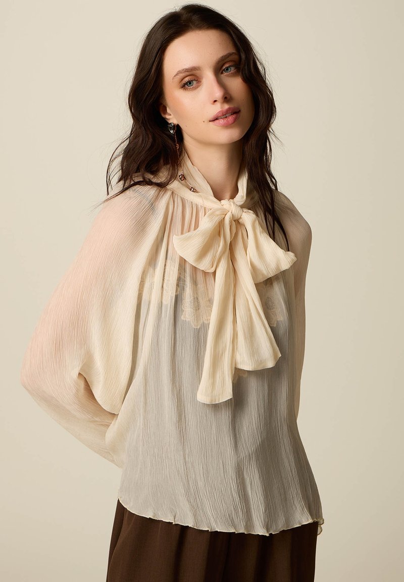 Młoda kobieta o długich ciemnych włosach, ubrana w przezroczystą kremową bluzkę z dużą kokardą przy kołnierzu oraz brązowe spodnie, na jednolitym beżowym tle.