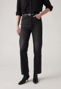 Zwarte high-waisted jeans met eenrechte snit, voorzien van voorzakken, een knoopsluiting en riemlussen, gecombineerd met zwarte enkellaarsjes.