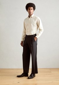 Camicia color beige chiaro con bottoni e maniche lunghe, abbinata a pantaloni neri su misura e scarpe nere, su uno sfondo tenue.