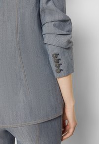 Blazer in denim grigio con finitura testurizzata, caratterizzato da maniche arrotolate e quattro bottoni decorativi sui polsini. Dettaglio cucito lungo le cuciture.