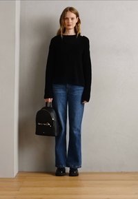 Pull à côtes noir, jean évasé bleu, mini sac à dos noir avec des éléments en métal argenté, et chaussures noires sur un sol en bois.