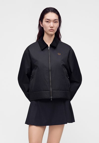 Femme aux longs cheveux foncés portant une veste noire zippée avec un petit logo doré et une jupe plissée noire sur un fond uni.